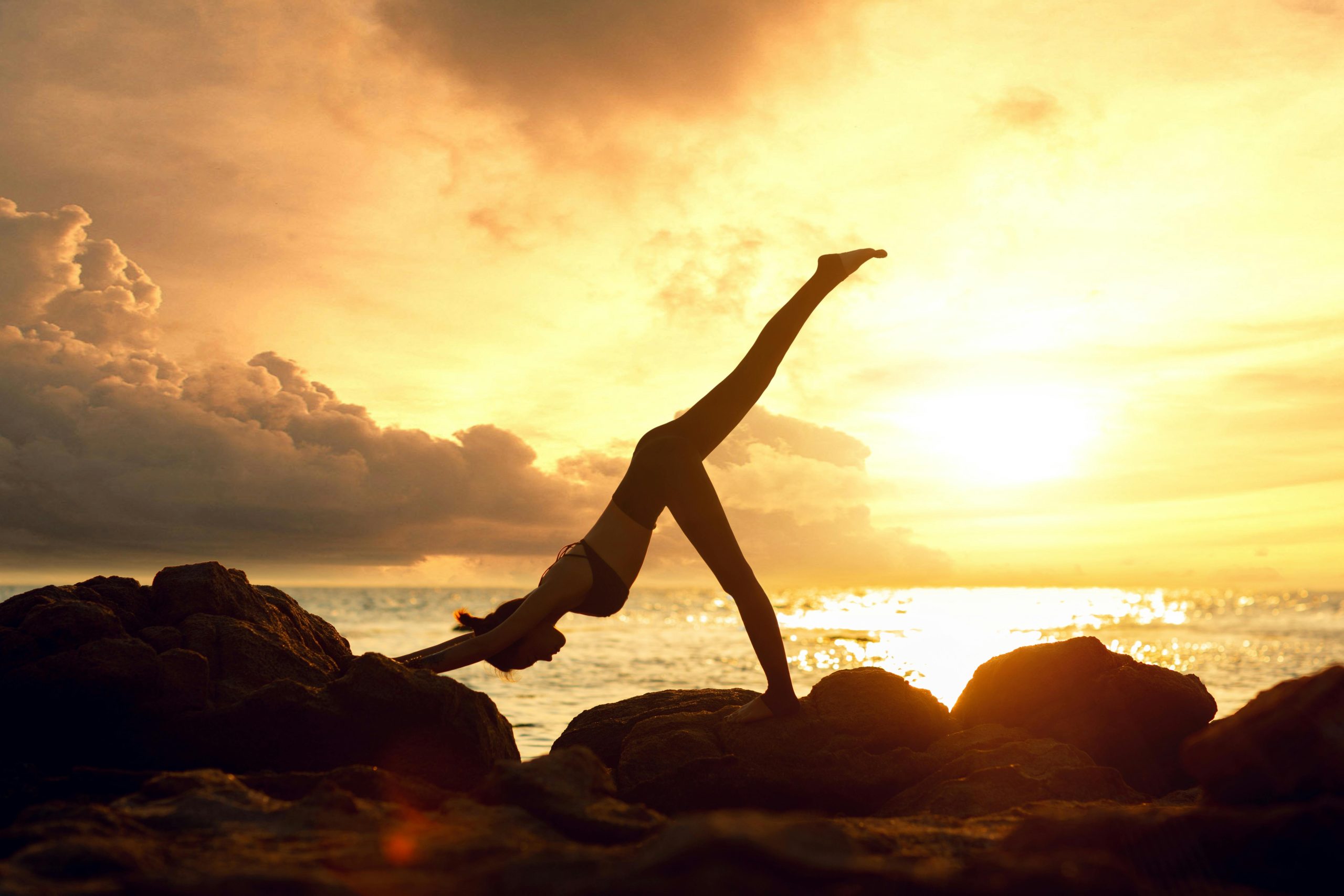 Silhouettes of two people on a rocky shoreline at sunset, arms raised toward the sky over the ocean. The scene is bathed in golden light.