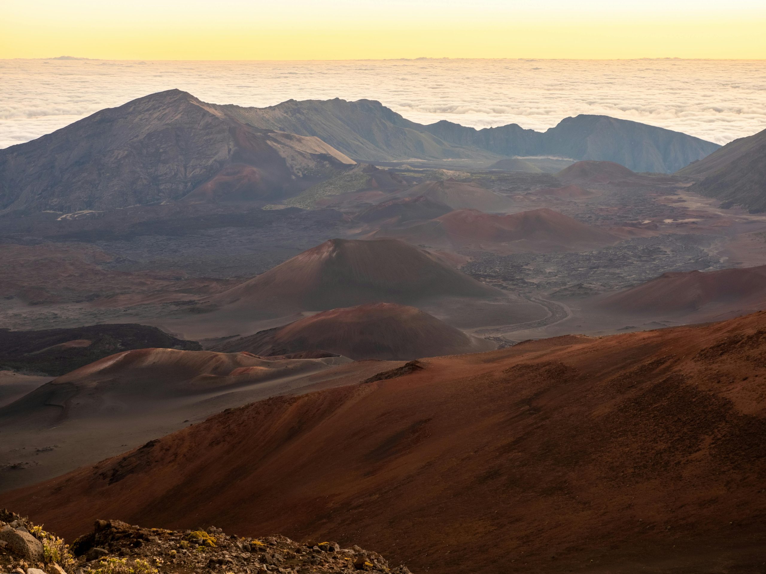 Volcanic landscape with reddish-brown cones, rugged mountains, and a valley beneath a pale cloud layer at sunrise.